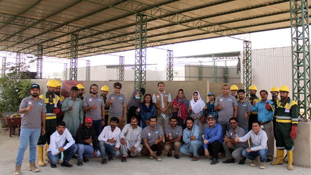 A large group of men and women, many in construction hats, stand posing for the camera under a high warehouse type roof.